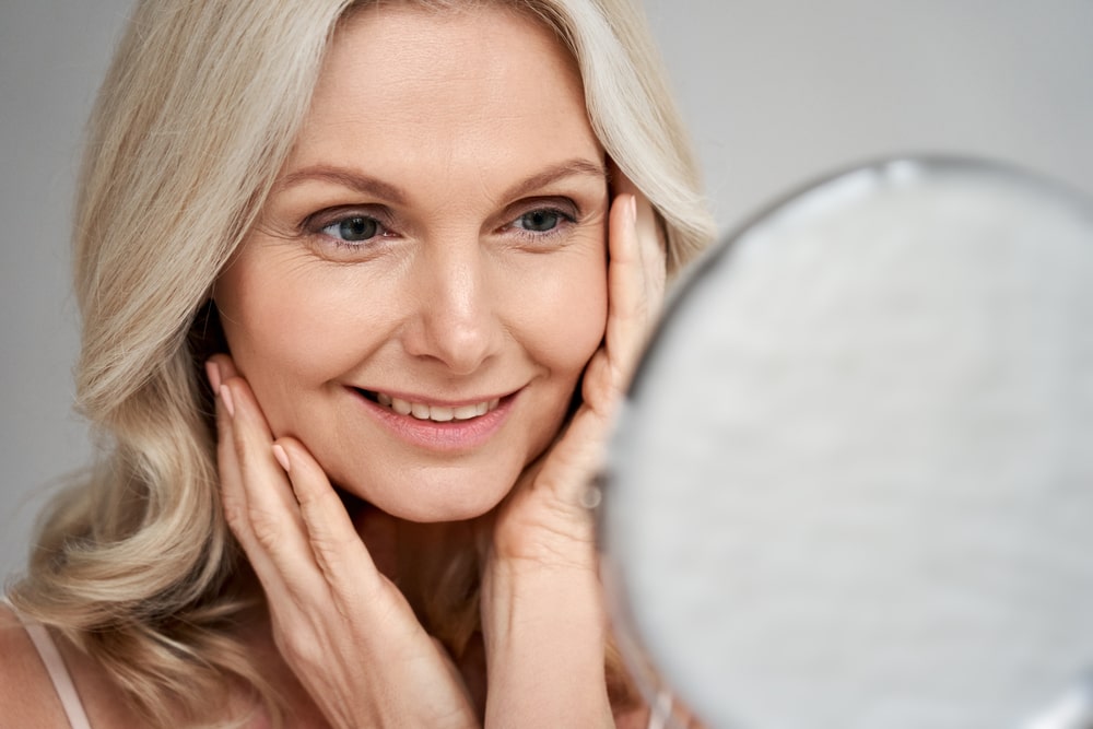 Happy 50s middle aged woman model touching face skin looking in mirror.